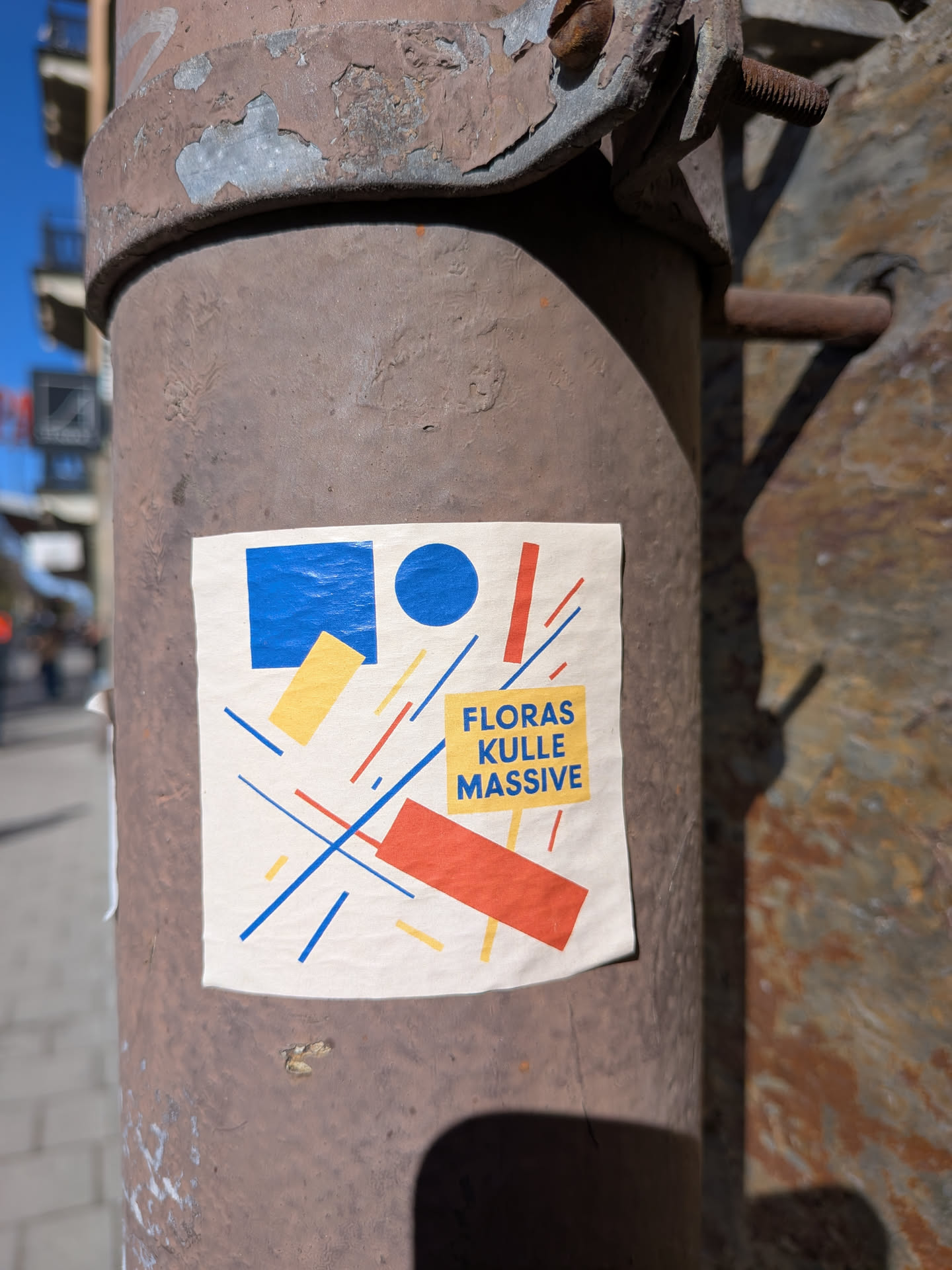 The sticker features an off-white background with a dynamic, abstract composition. In the upper left, there is a solid blue square and a solid blue circle. Below the blue square, a yellow horizontal rectangle is placed. In the lower right, a red horizontal rectangle is prominent. The design is crisscrossed by numerous thin, diagonal lines in blue and red, interspersed with shorter yellow line segments, creating a sense of movement. Centered on the right side, a yellow rectangular block contains the text "FLORAS KULLE MASSIVE" in bold blue capital letters. The overall aesthetic is reminiscent of De Stijl or Constructivist art, utilizing primary colors and basic geometric forms.