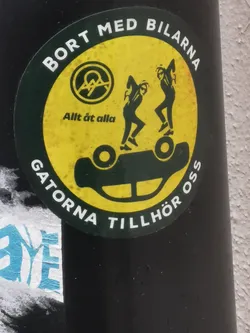 Street sticker A circular sticker with a dark green border and a vibrant yellow interior. The border features white uppercase text in Swedish. The central image on the yellow background shows two dynamic, dark green or black stylized figures dancing atop an overturned, dark green or black car. In the upper left of the yellow area, there's a dark green circular emblem containing a white stylized 'A' within a circle, a common anarchist symbol. Below this emblem, smaller dark green text is visible. The sticker exhibits signs of wear and tear, with some speckling and texture on the yellow surface.