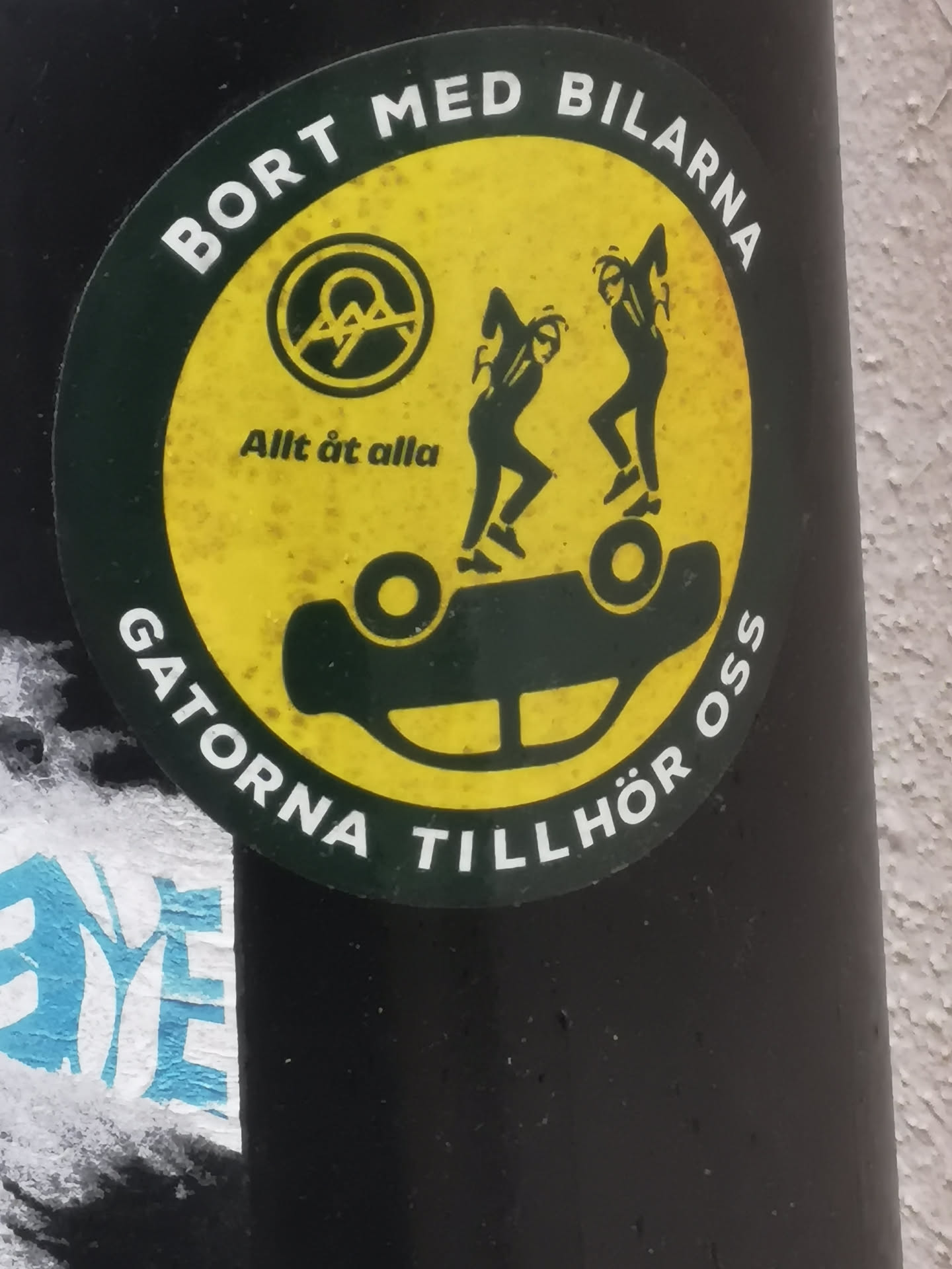 A circular sticker with a dark green border and a vibrant yellow interior. The border features white uppercase text in Swedish. The central image on the yellow background shows two dynamic, dark green or black stylized figures dancing atop an overturned, dark green or black car. In the upper left of the yellow area, there's a dark green circular emblem containing a white stylized 'A' within a circle, a common anarchist symbol. Below this emblem, smaller dark green text is visible. The sticker exhibits signs of wear and tear, with some speckling and texture on the yellow surface.