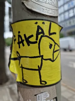 This image shows a yellow sticker affixed to a gray pole.  The sticker features a simple black line drawing of a dog, and the word "FÄKAL" (German for "fecal") is written above the drawing in black. The style of the drawing is reminiscent of a child's drawing.