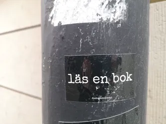 Street sticker The sticker is rectangular with a glossy black background. It features white, sans-serif text in two lines. The main text, 'läs en bok', is larger and centered. Below it, in smaller font, is the hashtag '#nobooksno party'. The sticker shows some reflections from the surrounding environment, indicating its glossy surface and physical presence on a textured, dark grey pole.