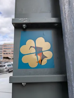 Street sticker A square blue sticker featuring a gold four-leaf clover made of four heart shapes. A thin blue cross divides the clover. At the center is a stylized, geometric bird graphic with black, white, orange, and pink details.