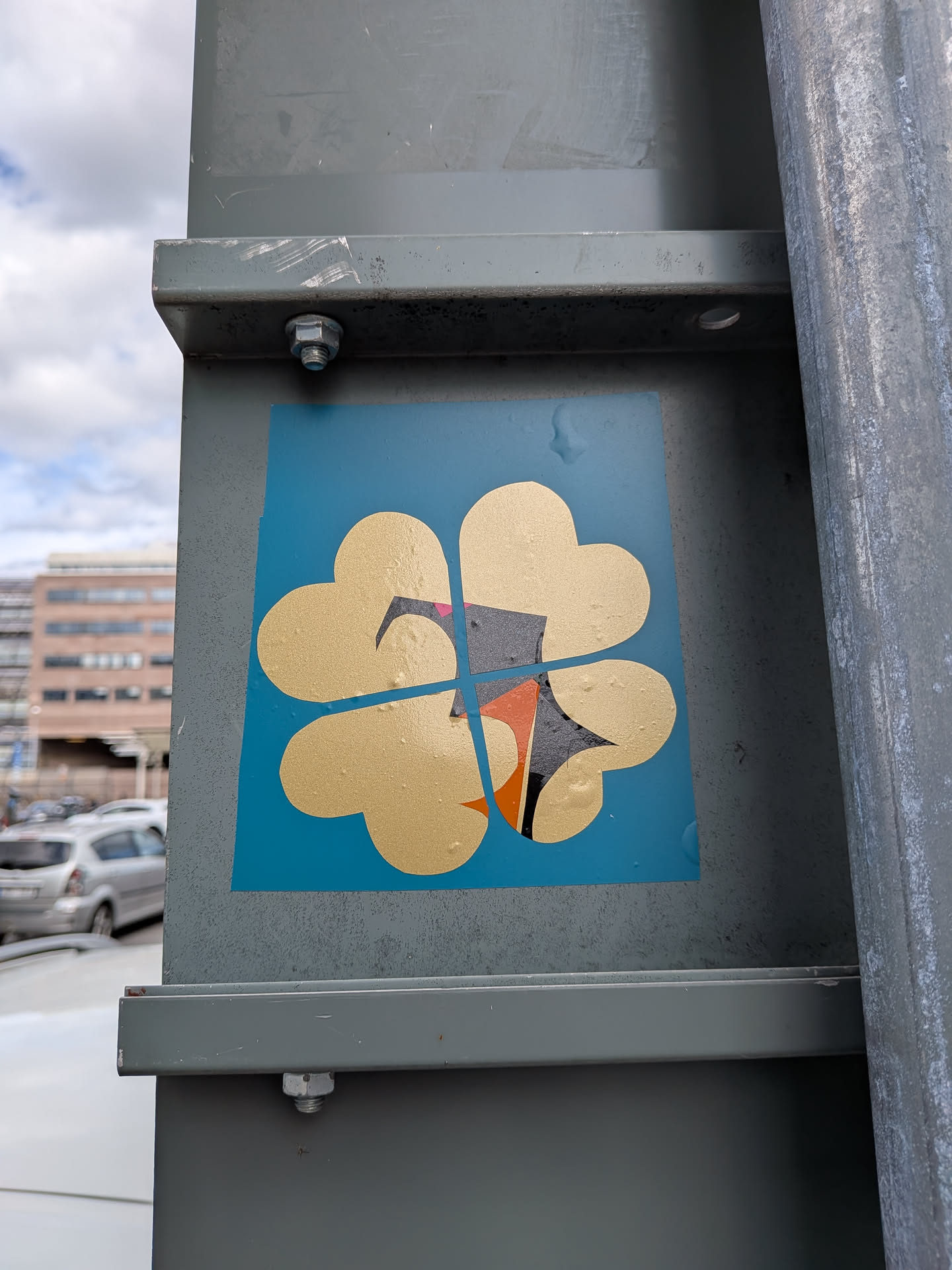 A square blue sticker featuring a gold four-leaf clover made of four heart shapes. A thin blue cross divides the clover. At the center is a stylized, geometric bird graphic with black, white, orange, and pink details.