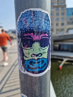 A vibrant sticker featuring a stylized monkey face, adorned with a patterned headwrap and sunglasses. The artwork is colorful and eye-catching, with a unique blend of textures and details.  The monkey's expression is cool and slightly mischievous. There are some small markings near the mouth that look like a signature or tag.