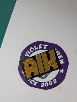 The sticker is composed of two layers. The base is a circular sticker with a deep purple background and a thin white outer border. White block letters arch around the top, spelling 'VIOLET CREW', with a white star on each side separating the words. At the bottom, also in white, it reads 'SINCE 2002'. Partially visible beneath the top layer, on the purple base, is a grey and white graphic that appears to be a stylized face or skull. Overlaid on this circular sticker is a smaller, rectangular black sticker. This top sticker prominently features the letters 'AIK' rendered in a distinctive retro-style, horizontally striped gold/yellow font. Small white starbursts or sparkles are scattered on the black background around the 'AIK' text.