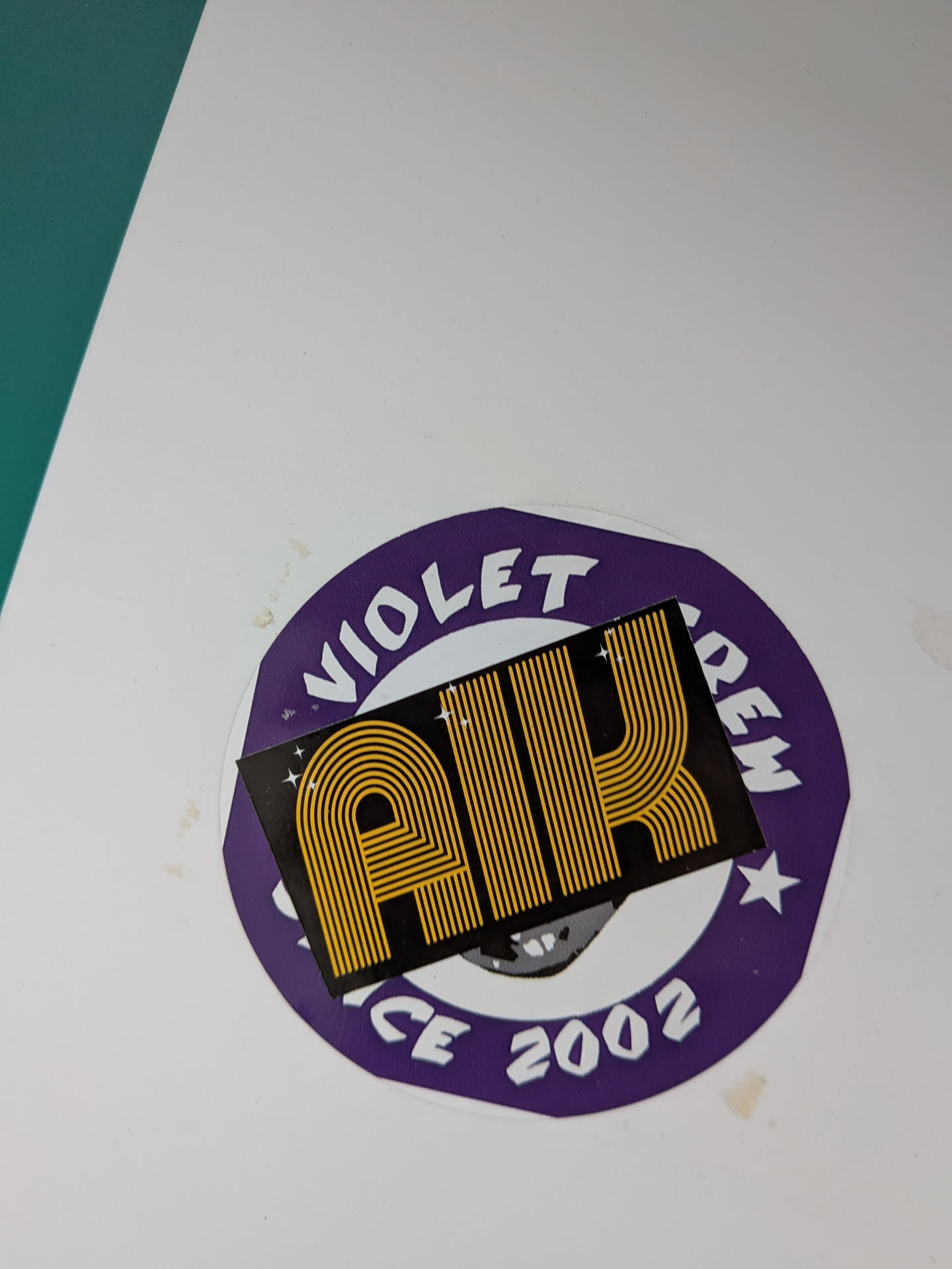The sticker is composed of two layers. The base is a circular sticker with a deep purple background and a thin white outer border. White block letters arch around the top, spelling 'VIOLET CREW', with a white star on each side separating the words. At the bottom, also in white, it reads 'SINCE 2002'. Partially visible beneath the top layer, on the purple base, is a grey and white graphic that appears to be a stylized face or skull. Overlaid on this circular sticker is a smaller, rectangular black sticker. This top sticker prominently features the letters 'AIK' rendered in a distinctive retro-style, horizontally striped gold/yellow font. Small white starbursts or sparkles are scattered on the black background around the 'AIK' text.