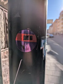 Street sticker A circular sticker featuring a prominent horizontal red band across its center. The background of the sticker, both above and below the red band, displays a shimmering, iridescent purple and pink holographic effect. Within the red band, there are stylized black characters that appear to read 'JMIII'. Below the red band, in the purple holographic section, there is a small, black, arrow-like or checkmark-shaped symbol pointing downwards and to the right. The sticker shows signs of wear, with some scratches and minor peeling around the edges, indicating it has been affixed for some time.