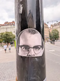 A dome-shaped black and white sticker depicting the upper half of a man's face. He wears thick-rimmed glasses and looks directly forward. The sticker has a white border and is pasted on a black pole.