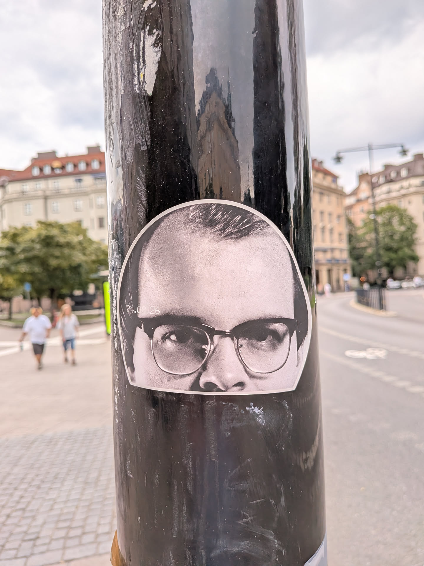 A dome-shaped black and white sticker depicting the upper half of a man's face. He wears thick-rimmed glasses and looks directly forward. The sticker has a white border and is pasted on a black pole.
