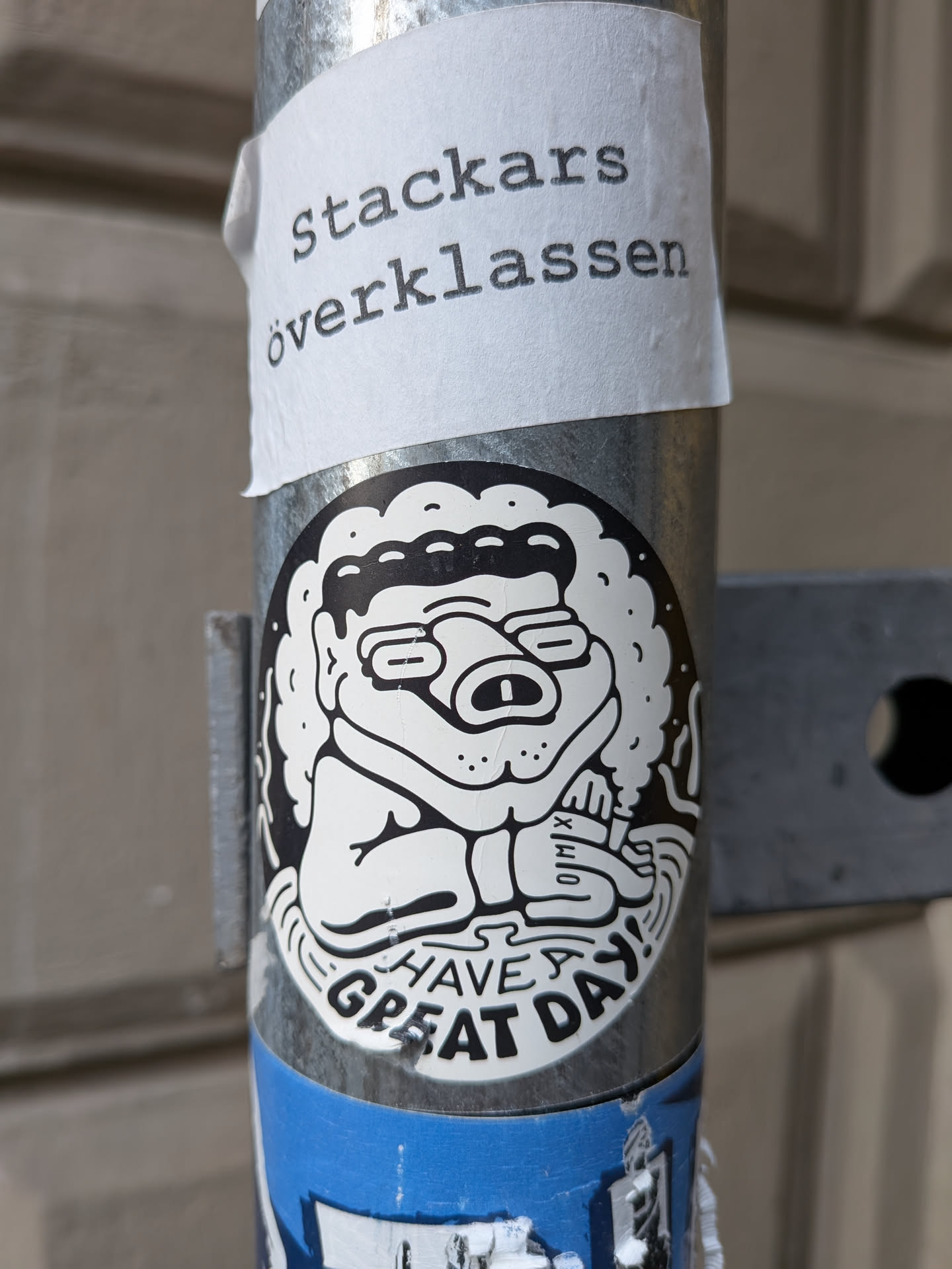 The sticker at the center of the image features a monochrome cartoon character with a distinctive, slightly grumpy-looking face. The character has round glasses, a prominent snout with nostrils, and a fluffy, cloud-like outline surrounding its head. It appears to be sitting and holding its arms crossed. Below the character, the words 'HAVE GREAT DAY!' are written in bold, stylized letters. The sticker has a black outline and is mostly white with black detailing, giving it a classic cartoon aesthetic. There is also a small, barely legible symbol on the character's arm. A paper sticker with Swedish text is placed above it.