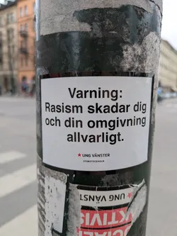 A white sticker on a dark pole. The sticker has black text in Swedish. The sticker states 'Varning: Rasism skadar dig och din omgivning allvarligt.' which means 'Warning: Racism harms you and your environment seriously.' The bottom of the sticker shows the logo and name 'UNG VÄNSTER STORSTOCKHOLM'.