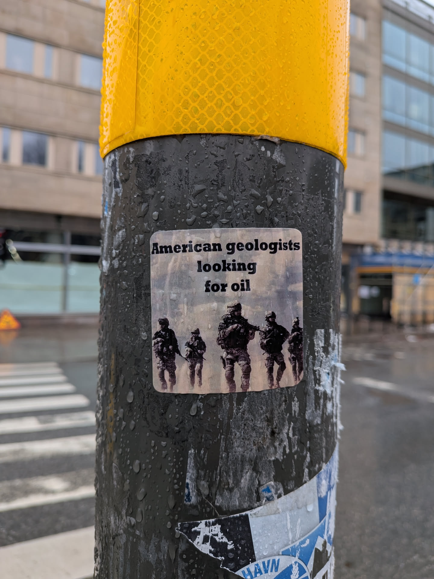 The sticker is a rectangular shape with slightly rounded corners, featuring a light grey or white background. At the top, in black sans-serif text, it reads 'American geologists looking for oil'. Below the text, there is an image of five figures dressed in military camouflage uniforms, each carrying what appears to be a rifle. They are depicted walking in a line from left to right across the sticker. The figures are rendered in a somewhat gritty, realistic style, with muted greens, browns, and greys for their uniforms. The background behind the soldiers is a light, hazy, almost sepia-toned landscape, possibly suggesting a desert or dusty environment. The sticker itself appears slightly worn and is covered in small water droplets, indicating it's outdoors on a wet day.