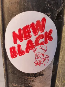 A round white sticker with red text and an illustration of a chef. The text says "NEW BLACK" in a bold, stylized font. The chef is depicted wearing a chef's hat and uniform, and is giving an okay sign.