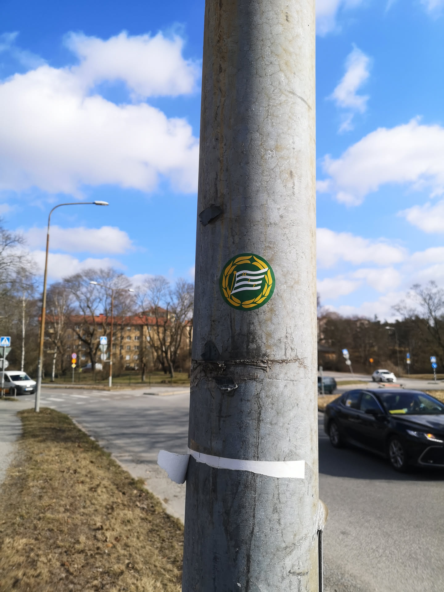 The sticker is circular, predominantly featuring shades of green, yellow, and white. It has a bright yellow border resembling a laurel wreath or a leafy garland. Inside this border, there is a dark green circular background. At the center of the green background, a stylized white flag or banner is depicted with three horizontal, wavy stripes, giving the impression of motion. The sticker appears to be in good condition, contrasting with the weathered, grey metal pole it is affixed to.