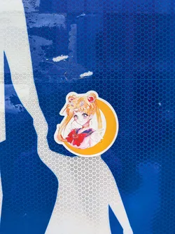 Street sticker A die-cut sticker featuring the anime character Sailor Moon (Usagi Tsukino). She is depicted from the chest up, with her signature blonde hair in odango buns, blue eyes, and wearing her sailor uniform with a prominent red bow. She is framed by a large, bright yellow-orange crescent moon. The sticker has a white border and is adhered to a blue textured reflective surface.
