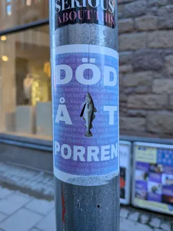 Street sticker The central sticker is predominantly light purple with large white lettering. The top part features the words 'SERIOUS ABOUT THIS' in white and pink on a black background. Below this, the sticker prominently displays the text 'DÖD ÅT PORREN' in large, bold, white sans-serif font. A graphic of a fish, resembling a cod, is depicted hanging from a thin string directly below the letter 'Å'. The fish is illustrated in grayscale with black details and appears to be wearing a small black tie. The light purple background has a repeating pattern of smaller text, mostly illegible, but appearing to be in Swedish, with phrases like 'SAMTYCKE ÄR INTE TILL SALU' (Consent is not for sale) faintly visible. The sticker has a slightly weathered appearance, consistent with being applied outdoors.
