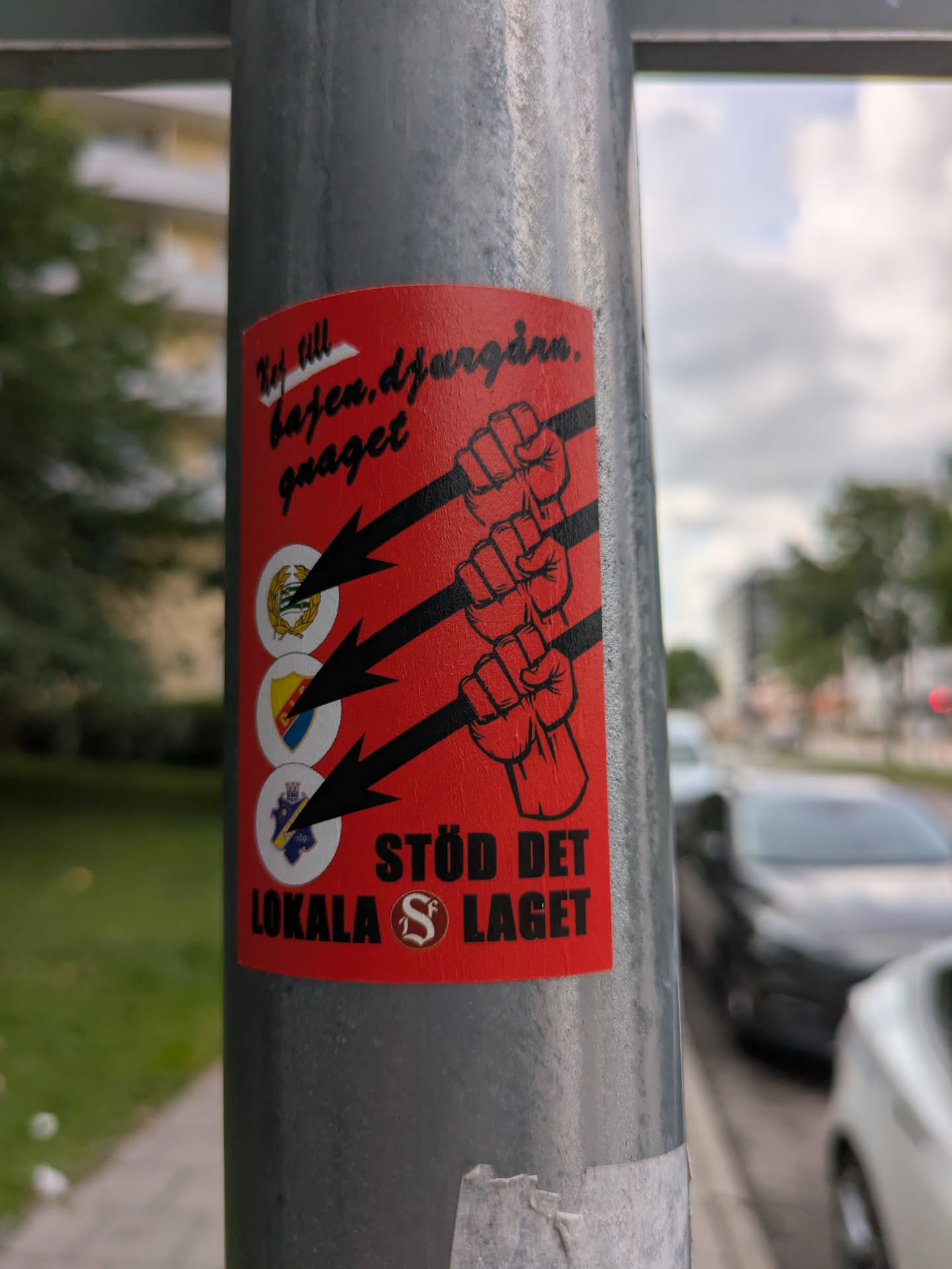 A rectangular red sticker affixed to a grey metal pole. The design features three black outlined fists stacked vertically on the right side, with three thick black arrows pointing towards them from the left. On the left side, corresponding to each arrow, are three circular white emblems, each containing the crest of a different Swedish football club: Hammarby IF (top, green wreath on yellow), Djurgårdens IF (middle, yellow and blue shield), and AIK (bottom, blue shield with white castle/tower). Black text in a handwritten style at the top left reads 'Nej till bajen.djurgårn. gnaget'. At the bottom right, bold black block letters read 'STÖD DET LOKALA' and below it 'S LAGET', with a stylized 'S' inside a circle between 'LOKALA' and 'LAGET'. The sticker has a slightly textured, worn appearance, indicating it is a physical object.
