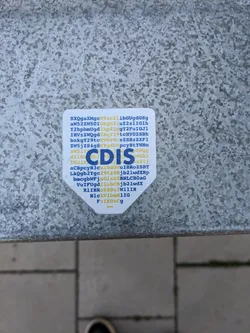 Street sticker A small, hexagon-shaped sticker with the letters 'CDIS' prominently displayed in the center. It is predominantly white with blue and yellow text. The sticker is affixed to a grey metallic surface.  The surrounding text appears to be an alphanumeric code. There are no social media links or QR codes visible.