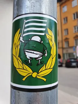 This sticker showcases a whimsical illustration of a rotund figure joyfully raising a flag with horizontal stripes.  The character is set against a vibrant green background, encircled by a cheerful yellow laurel wreath. The design is simple yet eye-catching.