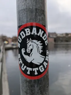 Street sticker A round sticker with a red border and black background featuring a stylized illustration of a muscular horse.  The text 'SÜDBANDE STUTTGART' is prominently displayed in a circular design around the horse. The sticker appears to be affixed to a metal pole near a body of water.
