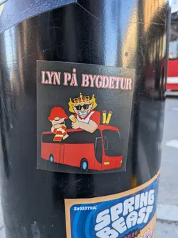 A rectangular sticker with a black background, featuring two cartoon characters riding in a red bus. The character on the left is smaller, resembling a gnome or dwarf, wearing a red pointed hat, a red and white striped scarf, and a red shirt. The character on the right is larger, depicted as a king or strongman with blonde hair, a golden crown, black sunglasses, and a red and white striped shirt, holding a yellow beer can. Behind the king character, three yellow objects resembling flares or pyrotechnics with red caps are visible. The red bus has light blue wheels and a front grille that suggests a Volvo model. White text with a red outline is prominently displayed at the top of the sticker.