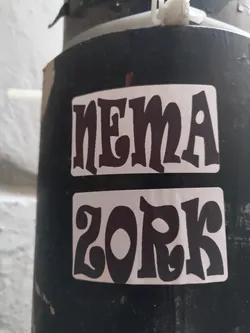 Two-part sticker featuring stylized text. The top sticker reads "NEMA" and the bottom sticker reads "ZORK". Both are in black text on a white background with a retro font style.  The stickers are affixed to a dark-colored pole outdoors.