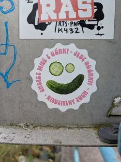 Street sticker The sticker is a white, scalloped-edge circle with a vibrant design. It depicts a cheerful face composed of two round cucumber slices for eyes and a whole green cucumber forming a wide smile. Around this central image, pink text is arranged in a circular pattern. The sticker is adhered to a textured, grey metallic surface.