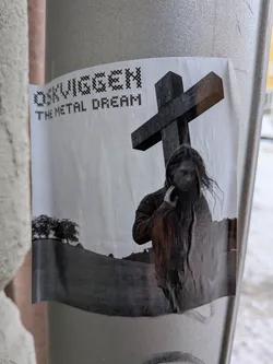 Street sticker A black and white sticker adhered to a light grey pole. The central image features a person with long, dark hair, looking downwards or away, with one hand near their face. Behind and to the right of the figure stands a large, dark, three-dimensional cross. In the background to the left, a rolling hill is visible with a cluster of trees at its crest. The overall scene is melancholic and stark. At the top left, the text "OSKVIGGEN" is written in a pixelated, cross-stitch-like font, with "THE METAL DREAM" directly below it in the same style. The sticker itself shows signs of wear, with slight wrinkles and peeling at the bottom edge.