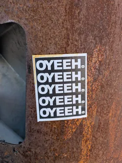 Street sticker The sticker is a vertical rectangle featuring a black background with white text and icons. The word "OYEEH." is printed five times in a bold, uppercase, sans-serif font, stacked vertically. To the immediate right of the period in each instance of "OYEEH.", there is a small, white skull icon. The sticker has a thin white border, and there is a noticeable yellowish-brown discoloration along its top and left edges, suggesting age or environmental exposure. It is affixed to a heavily rusted metal surface.