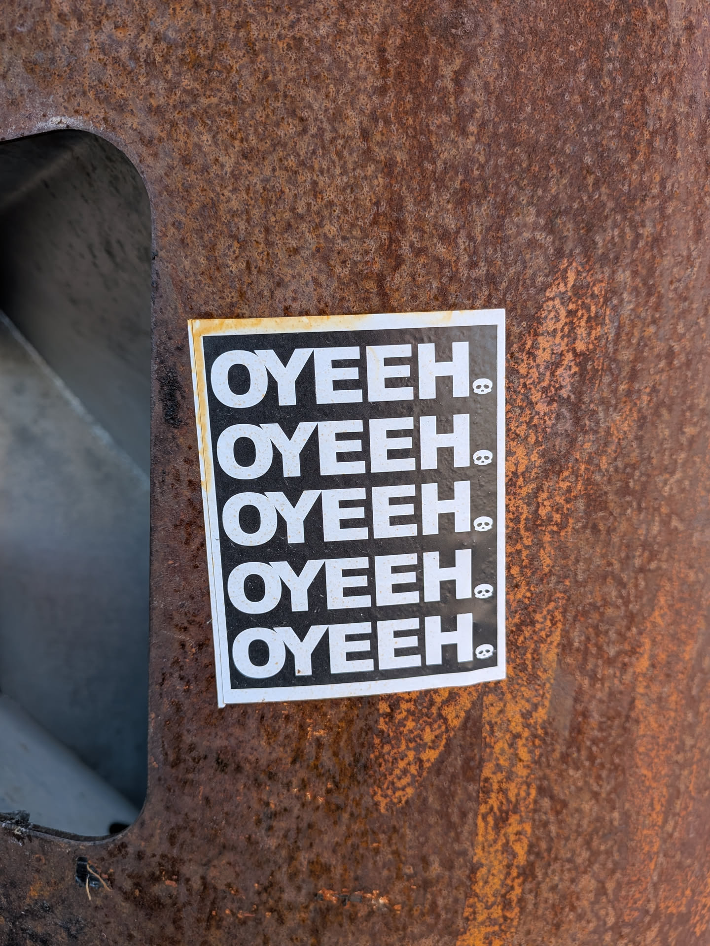 The sticker is a vertical rectangle featuring a black background with white text and icons. The word "OYEEH." is printed five times in a bold, uppercase, sans-serif font, stacked vertically. To the immediate right of the period in each instance of "OYEEH.", there is a small, white skull icon. The sticker has a thin white border, and there is a noticeable yellowish-brown discoloration along its top and left edges, suggesting age or environmental exposure. It is affixed to a heavily rusted metal surface.
