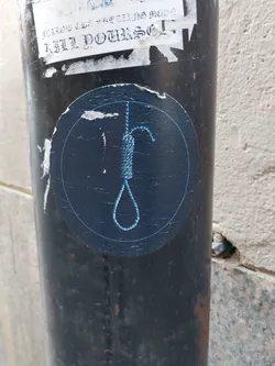 Street sticker A round dark blue sticker with a light blue outline depicts a noose. The sticker is affixed to a dark metal pole. Another sticker above it is partially visible, with some illegible text. The background is a light beige wall.