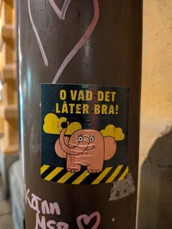 Street sticker The sticker is square-shaped with a dark blue/black background. In the center is a cartoonish pink elephant with large, round eyes, a wide smile, and rosy cheeks. It has its trunk raised and appears to be waving. Above the elephant are two fluffy yellow clouds. The bottom of the sticker features a pattern of diagonal yellow and black stripes, reminiscent of hazard tape. The text 'O VAD DET LÅTER BRA!' is prominently displayed in yellow, blocky letters at the top of the sticker.
