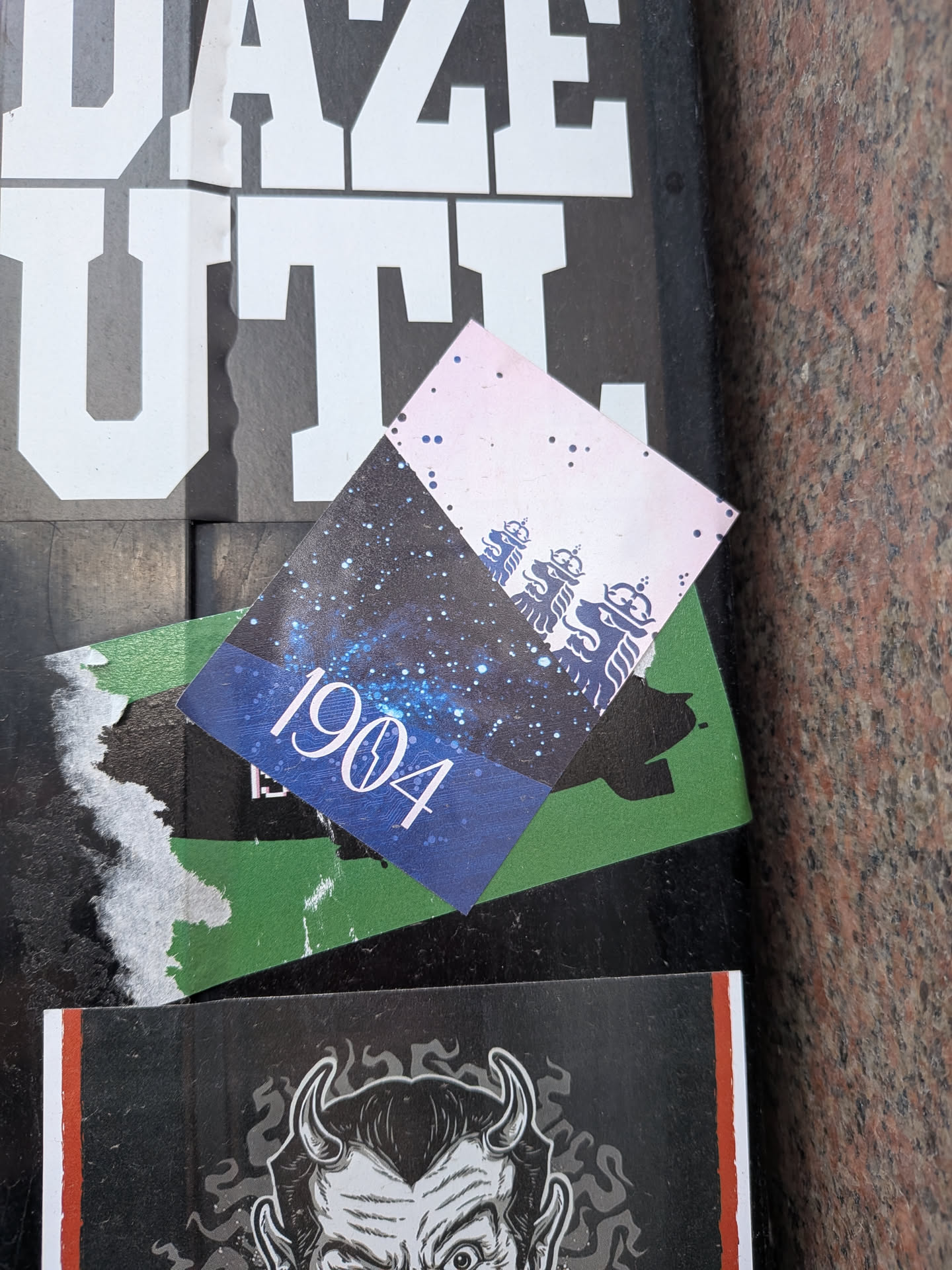 The central sticker is rectangular and oriented diagonally. It features a split design: the lower, larger section is a deep blue with a dense pattern of white and light blue speckles, evoking a starry night sky or galaxy. The number "1904" is prominently displayed in white, elegant serif font in the lower left of this section. The upper, smaller section is a pale pink or light purple, also with subtle dark speckles, and features a repeating pattern of three stylized, dark blue or purple crown-like symbols, possibly heraldic in nature. The sticker shows some signs of wear and is layered over a green sticker and partially under a larger sticker with white block letters.