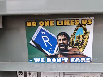 A sticker featuring Gerard Butler as Leonidas from 300, a periodic table element R, and a logo. The text on the sticker says: "NO ONE LIKES US - WE DON'T CARE"