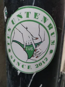 Street sticker A round sticker with a green border featuring an illustration of a woman's lower body and hands. The text "Tus Unternum since 2012" is written around the image.