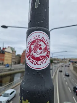 Street sticker A round sticker with a red background and white border. It features a black and white illustration of a person with a skull-like face, wearing a hat and holding a weapon. The text "Kamikaze Ultras Bordeaux" is written around the illustration.