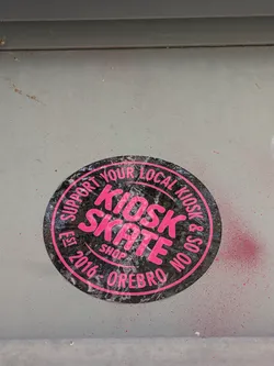 Street sticker A circular sticker with a black background and bright pink text. The text "KIOSK SKATE" is prominently displayed in large, bold, sans-serif font in the center. Around the edge, in a circular arrangement, are the words "SUPPORT YOUR LOCAL KIOSK & SOON EST. 2016 - OREBRO SHOP". The sticker appears to have a slightly textured or glossy finish, and there are some minor scuffs and imperfections visible, suggesting it is indeed a physical sticker. The background of the sticker has a subtle pattern that resembles a distressed texture.