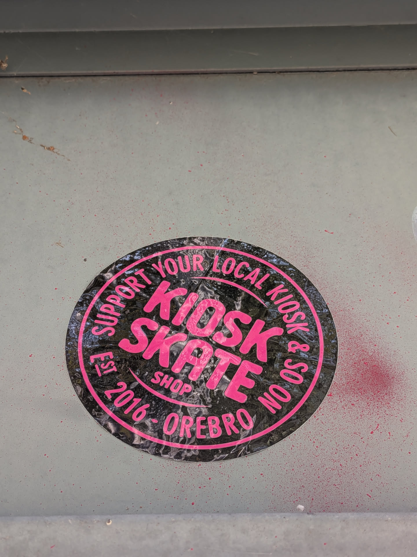 A circular sticker with a black background and bright pink text. The text "KIOSK SKATE" is prominently displayed in large, bold, sans-serif font in the center. Around the edge, in a circular arrangement, are the words "SUPPORT YOUR LOCAL KIOSK & SOON EST. 2016 - OREBRO SHOP". The sticker appears to have a slightly textured or glossy finish, and there are some minor scuffs and imperfections visible, suggesting it is indeed a physical sticker. The background of the sticker has a subtle pattern that resembles a distressed texture.