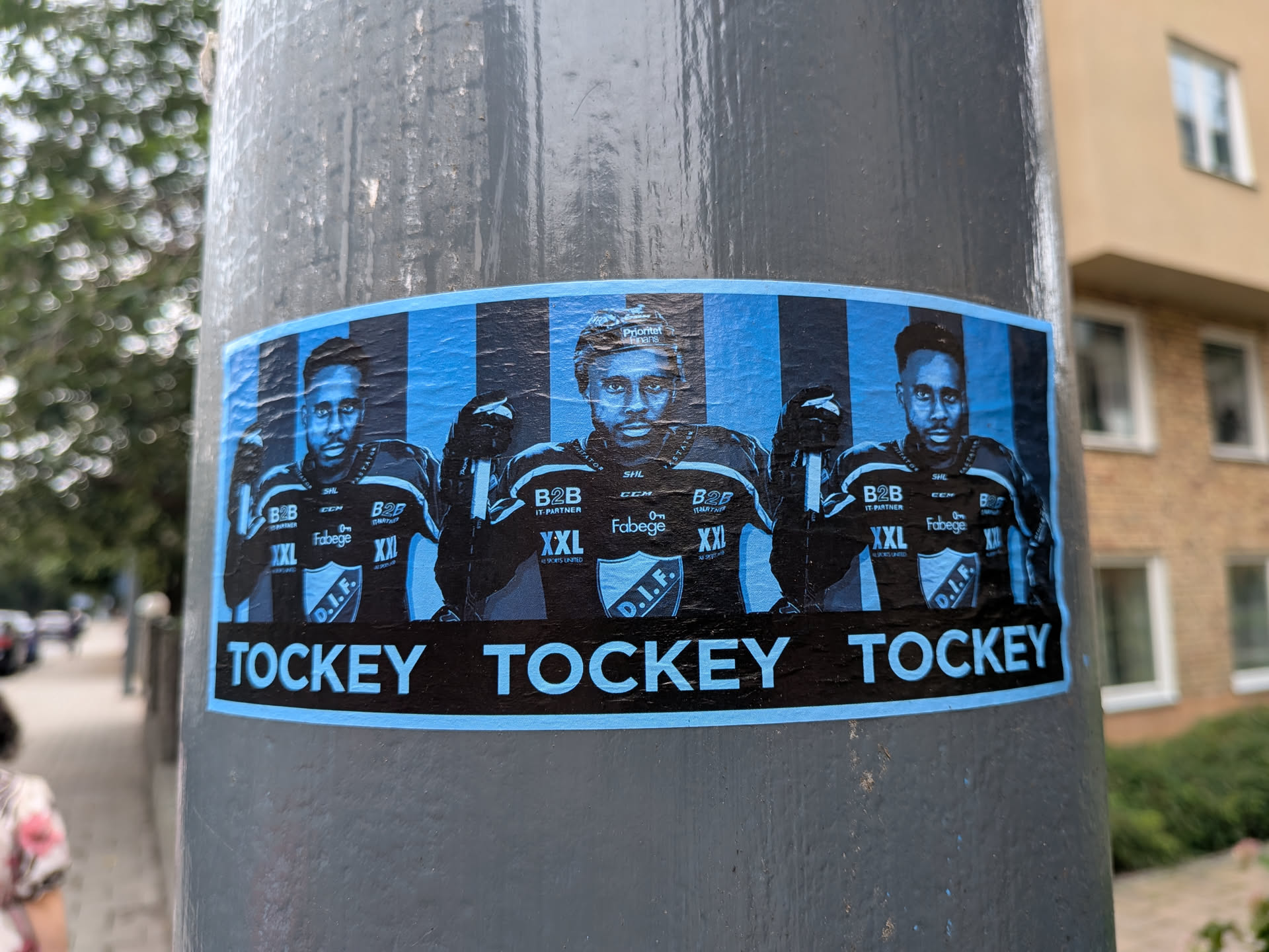 The sticker is centered on a grey, cylindrical pole and features a repeated image of three hockey players. The players are depicted from the chest up, wearing hockey jerseys and helmets, and are rendered in a monochromatic blue and black style against a striped blue background. The jerseys have various sponsor logos, including 'B2B IT-PARTNER', 'CCM', 'Fabeg e', and a crest with 'D.I.F.'. Below the players, the word 'TOCKEY' is prominently displayed in large, white, sans-serif letters on a black background, which is bordered by a thin blue line. The sticker shows some signs of wear and creasing, indicating it has been applied outdoors.