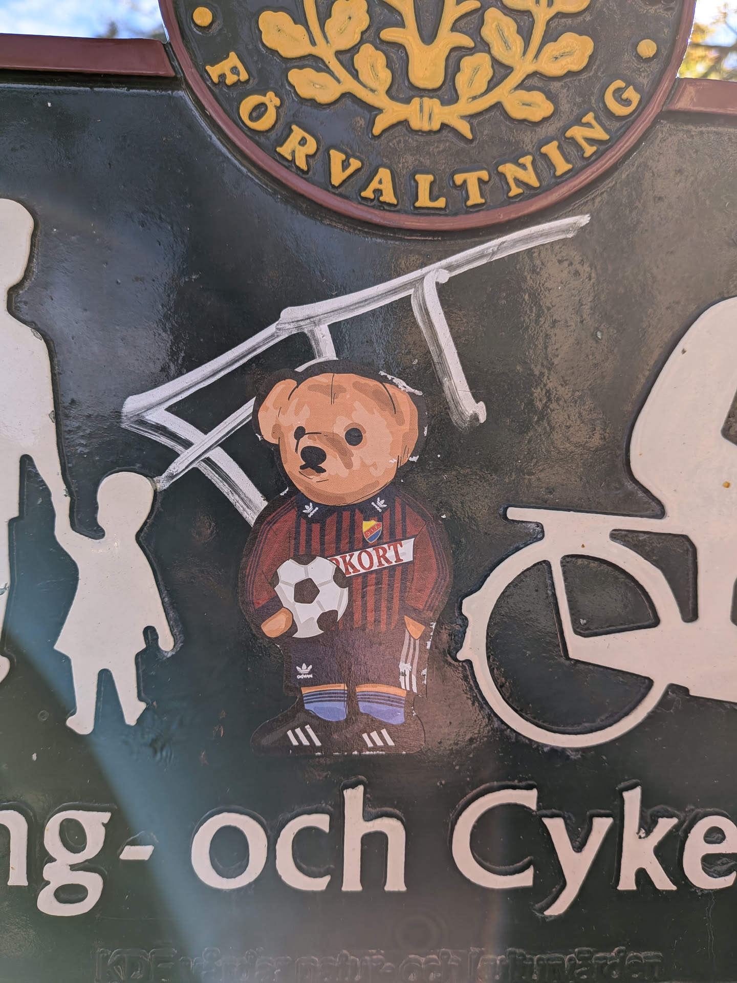 The sticker features a cartoonish teddy bear wearing a red and dark striped football jersey with yellow accents and the word 'KORT' in white letters. The jersey also has the Adidas logo and what appears to be a team crest. The bear is holding a white and black soccer ball and is wearing Adidas-branded soccer cleats and striped socks. The sticker is placed on a dark, textured surface, possibly a sign, and is slightly peeling at the edges.