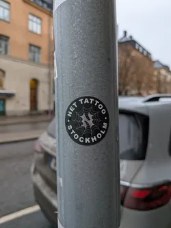 Street sticker A circular, black sticker with white text and a white spiderweb design in the center. The text 'S. NET TATTOO' is arranged in an arc at the top, and 'STOCKHOLM' is arranged in an arc at the bottom, separated by dots. The spiderweb has a central point with radiating lines and connecting strands, with a few small circular embellishments.