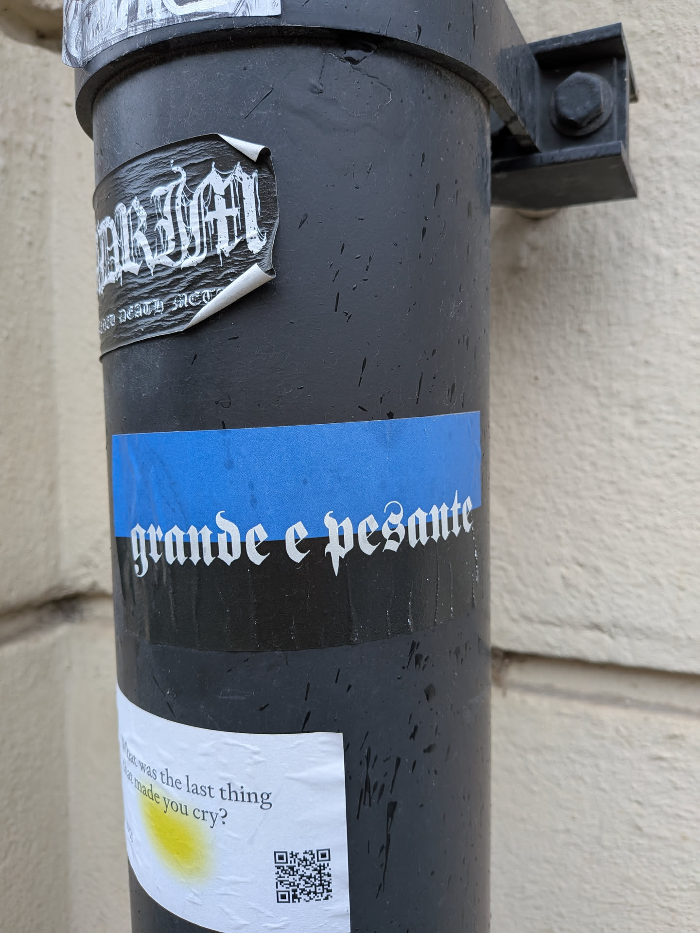 A rectangular sticker featuring a prominent horizontal blue band across its upper half, set against a black background. White text in a distinctive gothic or blackletter font reads 'grande e pesante'. The sticker shows some wear, minor bubbling, and is adhered to a textured, wet surface.