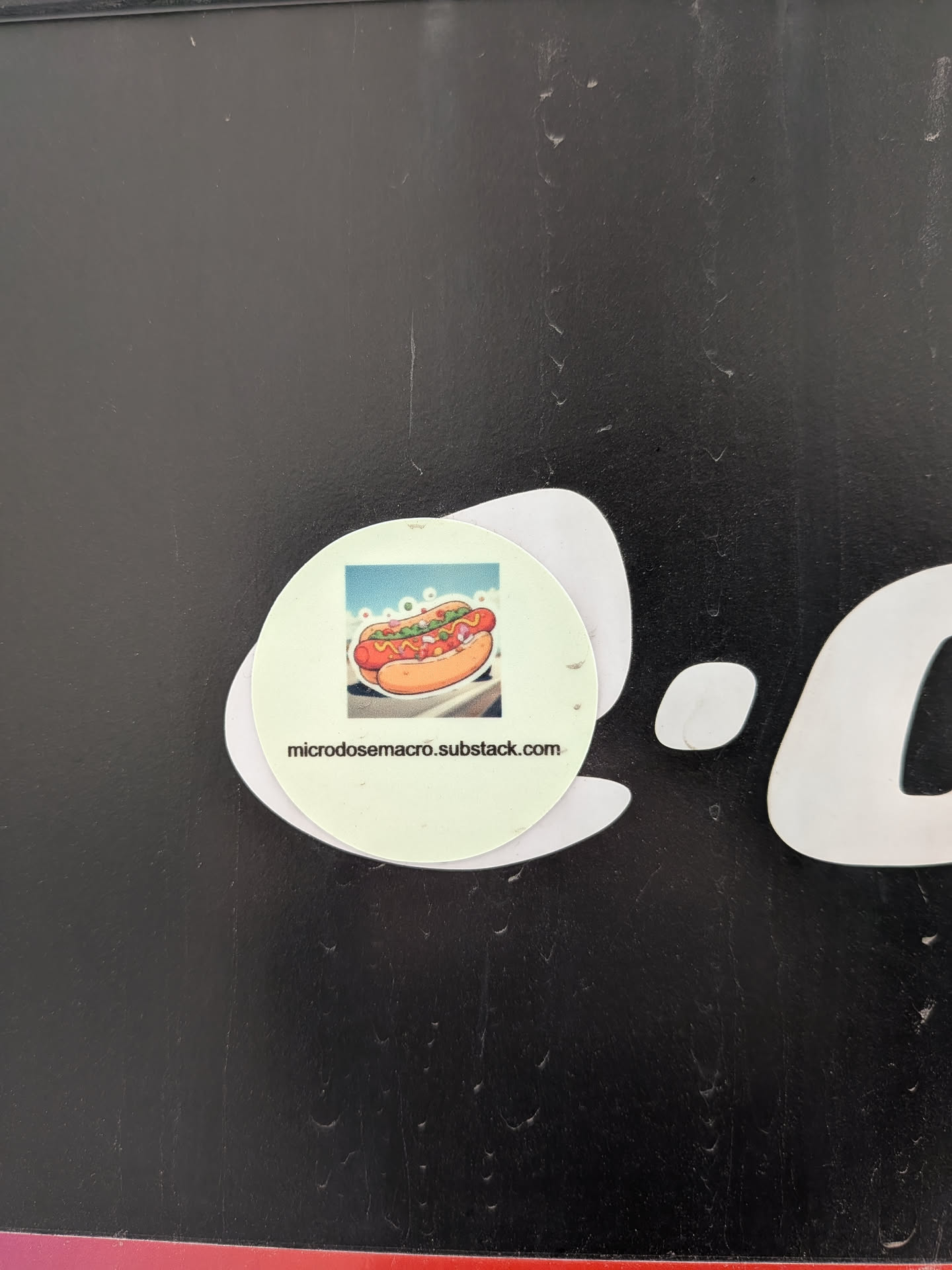A circular sticker with a light green background featuring a vibrant, cartoon-style illustration of a hot dog. The hot dog is reddish-brown in a golden-brown bun, generously topped with yellow mustard, green relish, and what appears to be chopped white onions. The background of the illustration shows a light blue sky with white, cloud-like shapes and a hint of a road or surface below. Below the illustration, in black sans-serif text, is the URL 'microdosemacro.substack.com'. The sticker is adhered to a dark, textured surface, with a larger, irregularly shaped white sticker partially visible beneath it.