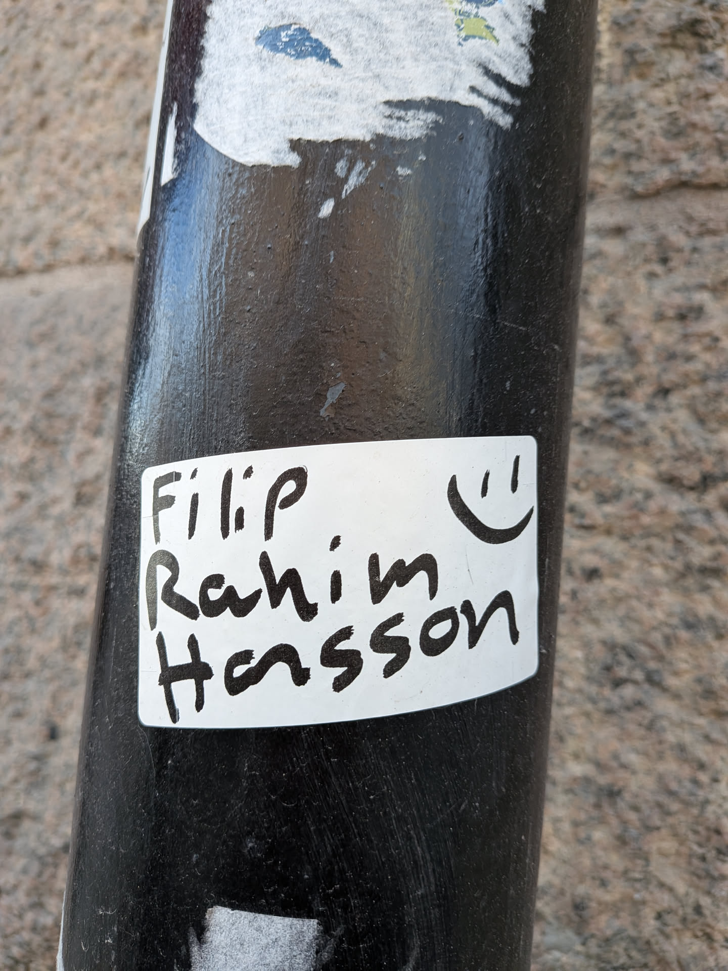 The sticker is a rectangular white label with slightly rounded corners, adhered to a dark, cylindrical pole. The surface of the sticker shows minor wear and conforms to the pole's curvature. Handwritten in bold black marker are the names 'Filip', 'Rahim', and 'Hasson', stacked vertically. To the right of the names, a simple black smiley face emoticon is drawn. The overall appearance is clean, personal, and minimalist.