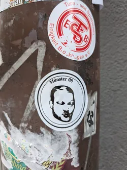 Street sticker A circular sticker featuring a minimalist, black-and-white portrait of a man's face. The face is stylized with bold black lines and white negative space, depicting a bald or closely shaven head with a beard or stubble. Above the portrait, curved along the top edge of the inner circle, is the text 'Münster 08' in black sans-serif font. The sticker has a white background with a thin black outer border.
