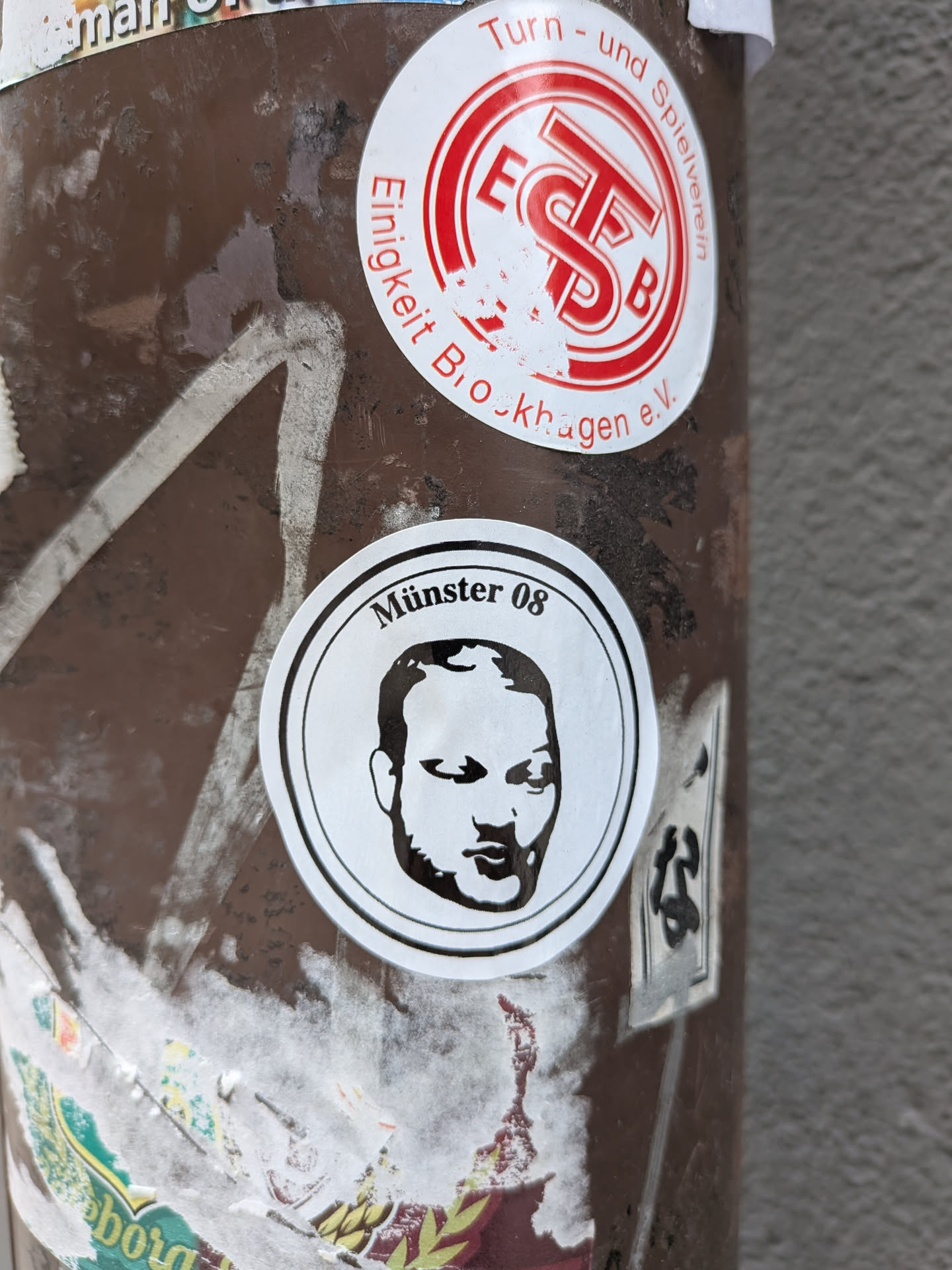 A circular sticker featuring a minimalist, black-and-white portrait of a man's face. The face is stylized with bold black lines and white negative space, depicting a bald or closely shaven head with a beard or stubble. Above the portrait, curved along the top edge of the inner circle, is the text 'Münster 08' in black sans-serif font. The sticker has a white background with a thin black outer border.