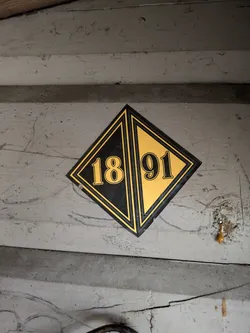 Street sticker The image features a diamond-shaped sticker centrally placed on a light grey, worn, possibly wooden surface. The sticker is divided diagonally, with the top-left half being black and the bottom-right half being a golden yellow. The number '18' is prominently displayed in golden yellow on the black half, and '91' is in black on the golden yellow half, completing the year '1891'. The sticker has a double border, an inner black line and an outer golden yellow line. The background surface shows signs of age, including cracks, dirt, and scuff marks, suggesting a historical or aged context for the sticker.