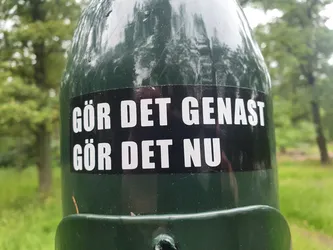 A black and white sticker on a green pole, with Swedish text that translates to "Do it immediately", "Do it now".