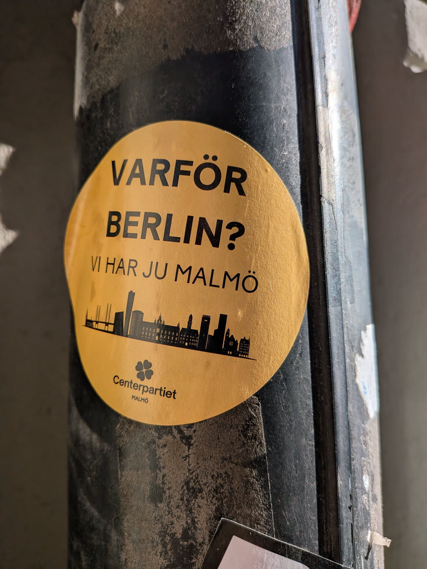 The sticker is circular with a golden-yellow background. It features bold black text at the top, asking 'VARFÖR BERLIN?' (Why Berlin?) and stating 'VI HAR JU MALMÖ' (We have Malmö). Below the text, there is a black silhouette of a cityscape, depicting various buildings and a bridge, representing Malmö. At the bottom, a small black four-leaf clover logo is present, followed by the text 'Centerpartiet' and 'MALMÖ' in smaller black font. The sticker appears slightly worn and dusty, consistent with outdoor placement.