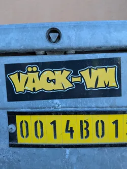 Street sticker A rectangular sticker with a solid black background, featuring the text 'VÄCK-VM' in a vibrant yellow, bubble-letter style. Each letter is outlined with a thick white border, which is then further outlined by a thin black line. Small white dots are scattered within the yellow fill of some letters, giving a textured or spray-paint effect. The two dots above the 'Ä' are rendered as distinct yellow circles with white outlines, resembling eyes and giving the letter a character-like appearance. The sticker is adhered to a textured, grey metallic surface.